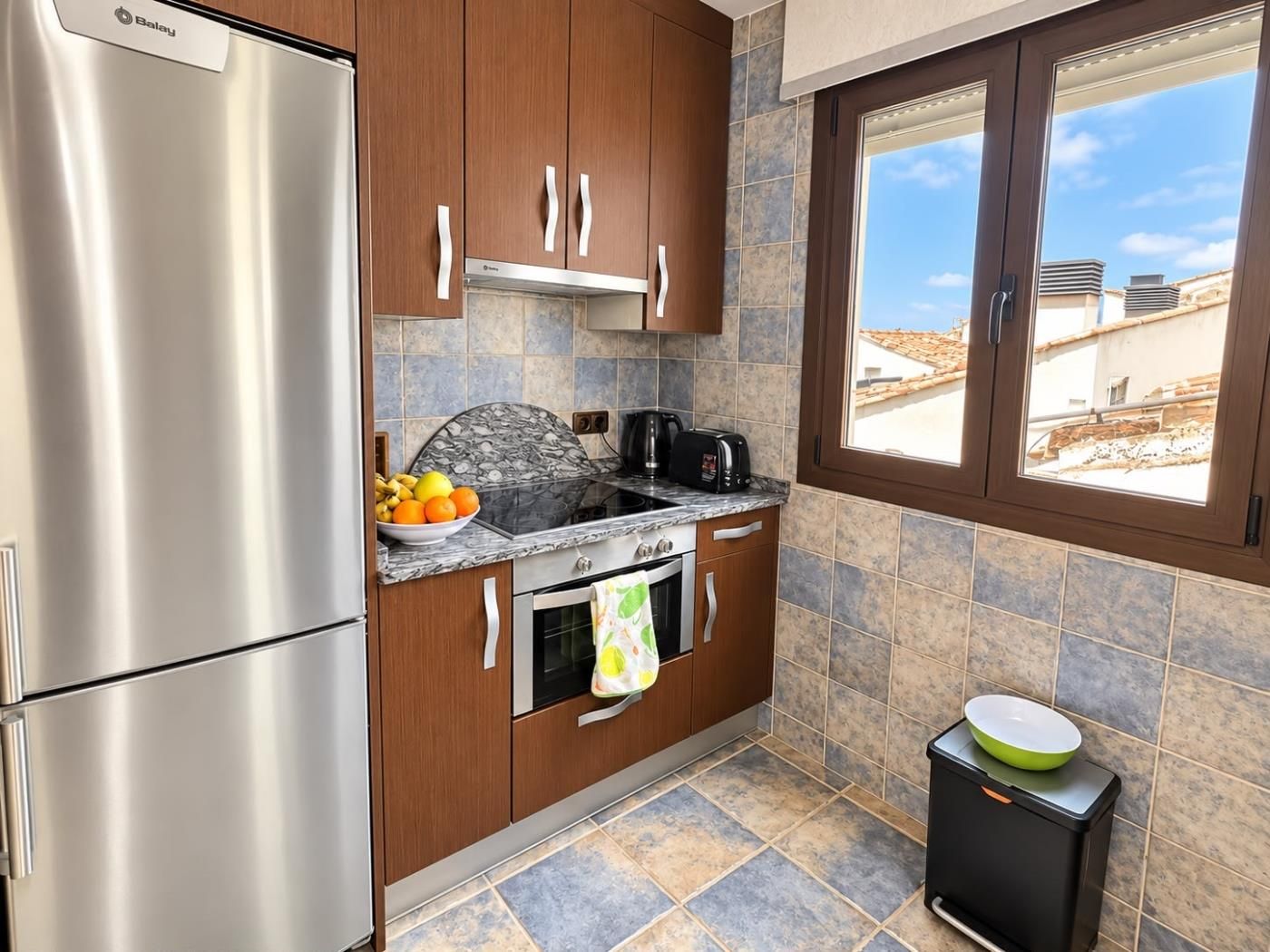A compact kitchen with a stainless steel refrigerator, wooden cabinets, a gas stove, and a window with a view of buildings.