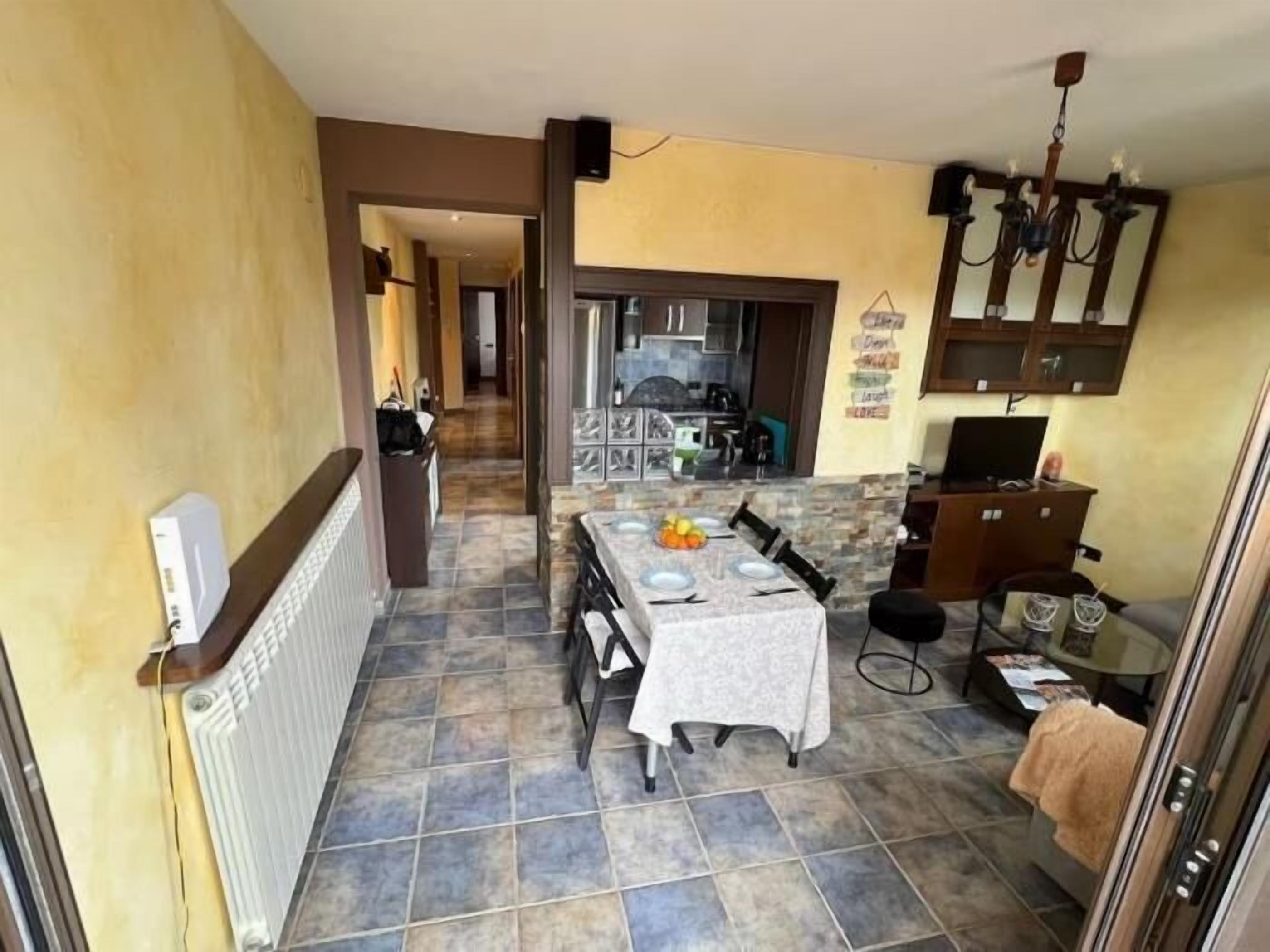 Un pasillo estrecho que lleva a un comedor con una mesa lista para dos personas, una cocina al fondo y un pequeño rincón de descanso con una silla y una mesa.