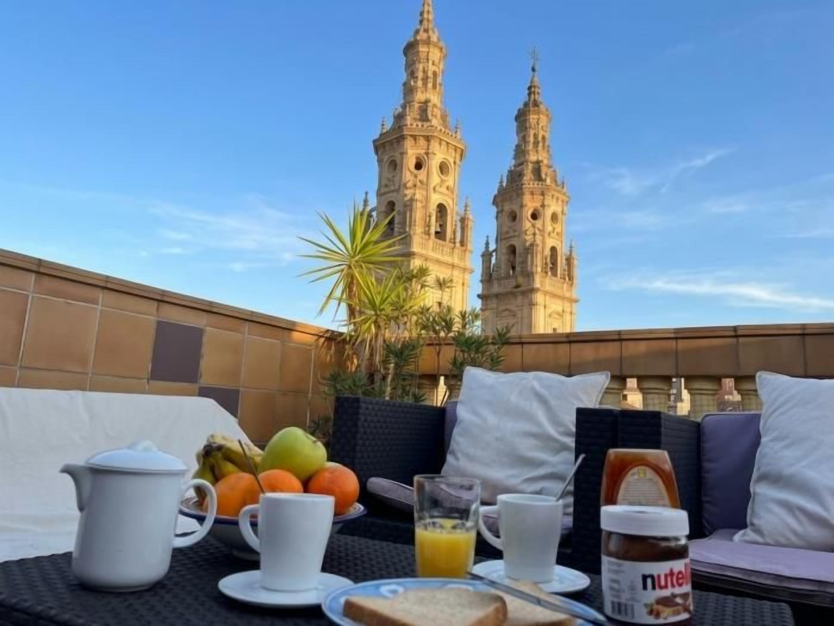 A breakfast setup with a view of two historic towers, a table with breakfast items, and a seating area with cushions.