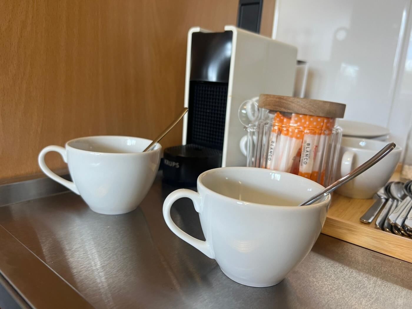 Dos tazas blancas con cucharas, una cafetera y un recipiente con lunares naranja y blanco.