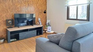 Een moderne woonkamer met een flatscreen televisie, een grijze bank, een houten accentmuur en een raam met zonwering.