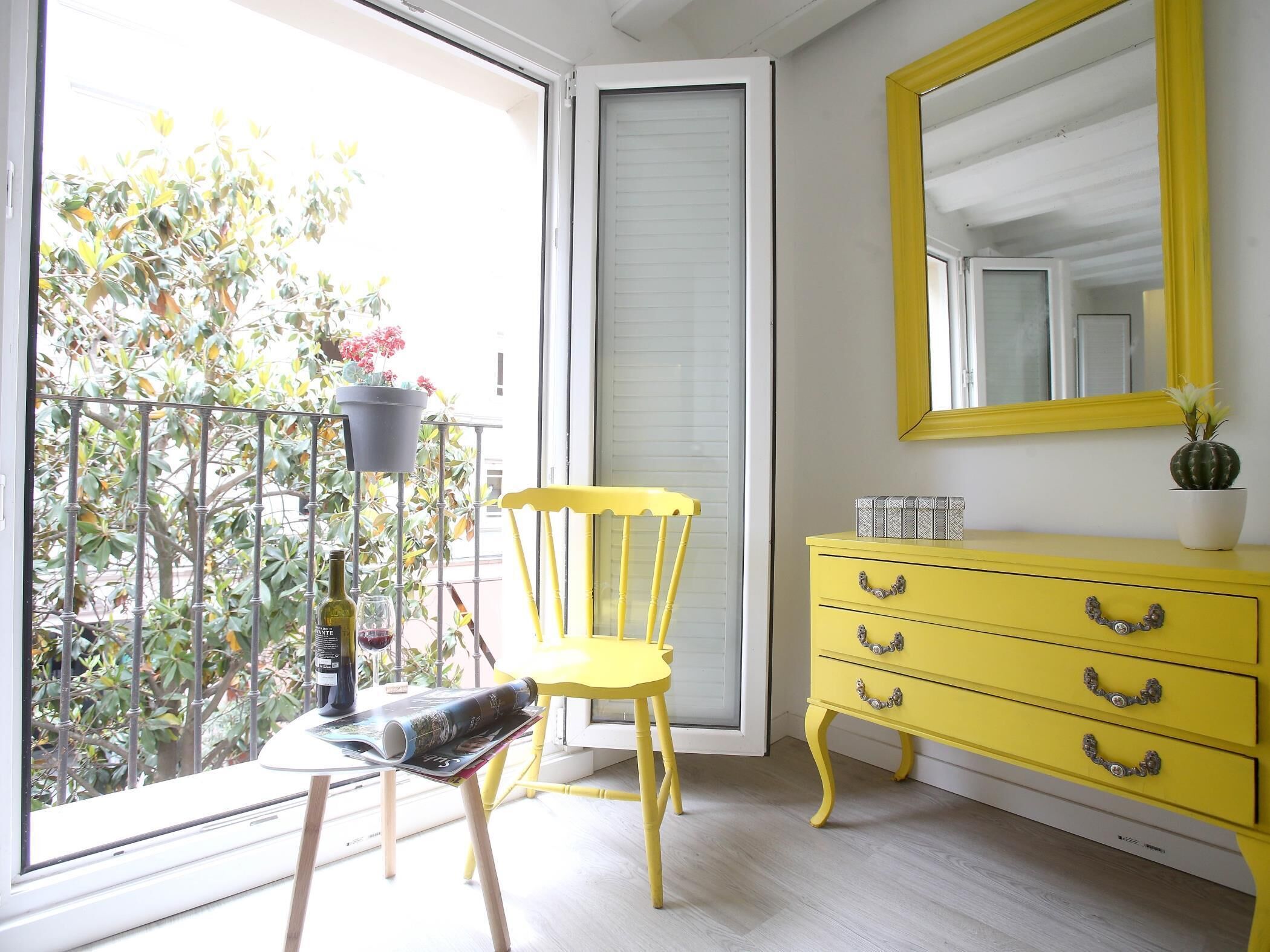 A room with a yellow dresser, a chair, a table with a magazine, a potted plant, and a balcony with plants and a railing.