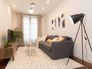 Una sala moderna con un sofá gris, una lámpara de pie, un televisor sobre un soporte de madera y una planta en maceta.