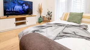 Makuuhuoneessa on sänky, jalustalla oleva televisio, pieni pöytä, jossa on kasvi, ja ikkuna, jossa on ohuet verhot.