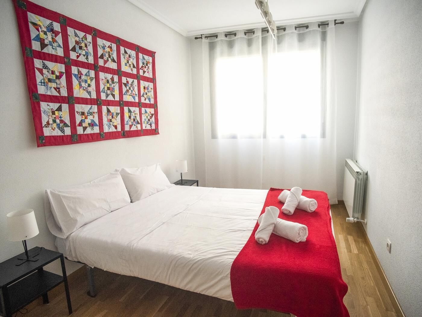 A hotel room with a bed, a red blanket, white pillows, a nightstand, a lamp, a quilt, and a window with curtains.