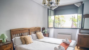Una cama doble con ropa blanca, un cabecero de madera, dos mesitas de noche con lámparas, una ventana con vistas a la vegetación y un radiador.