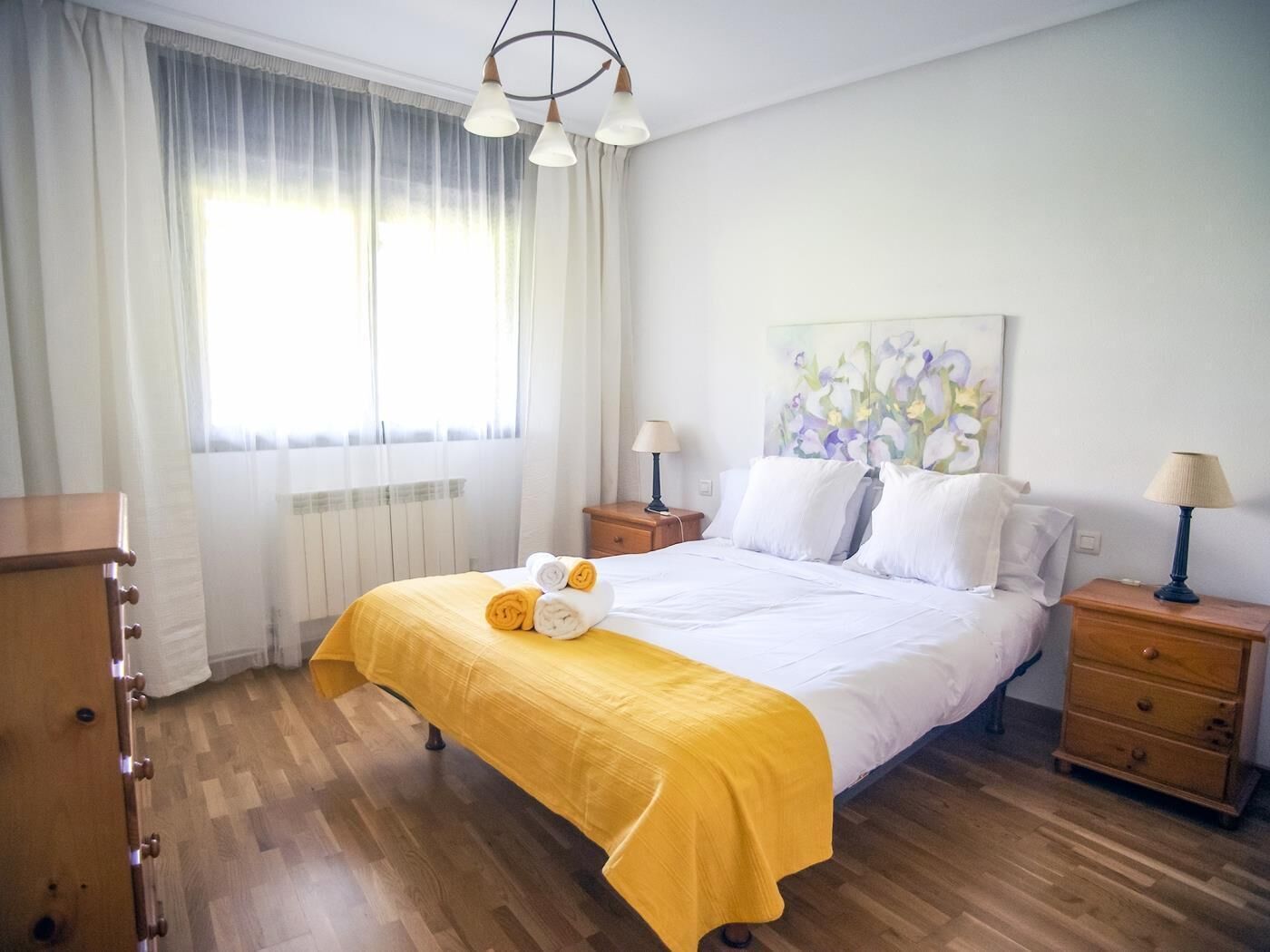 A hotel room with a bed, two bedside tables, a wooden dresser, a window with sheer curtains, and a painting on the wall.