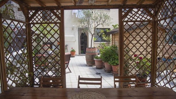 Una pérgola de madera con una mesa, sillas, plantas en macetas y una vista de un edificio al fondo.