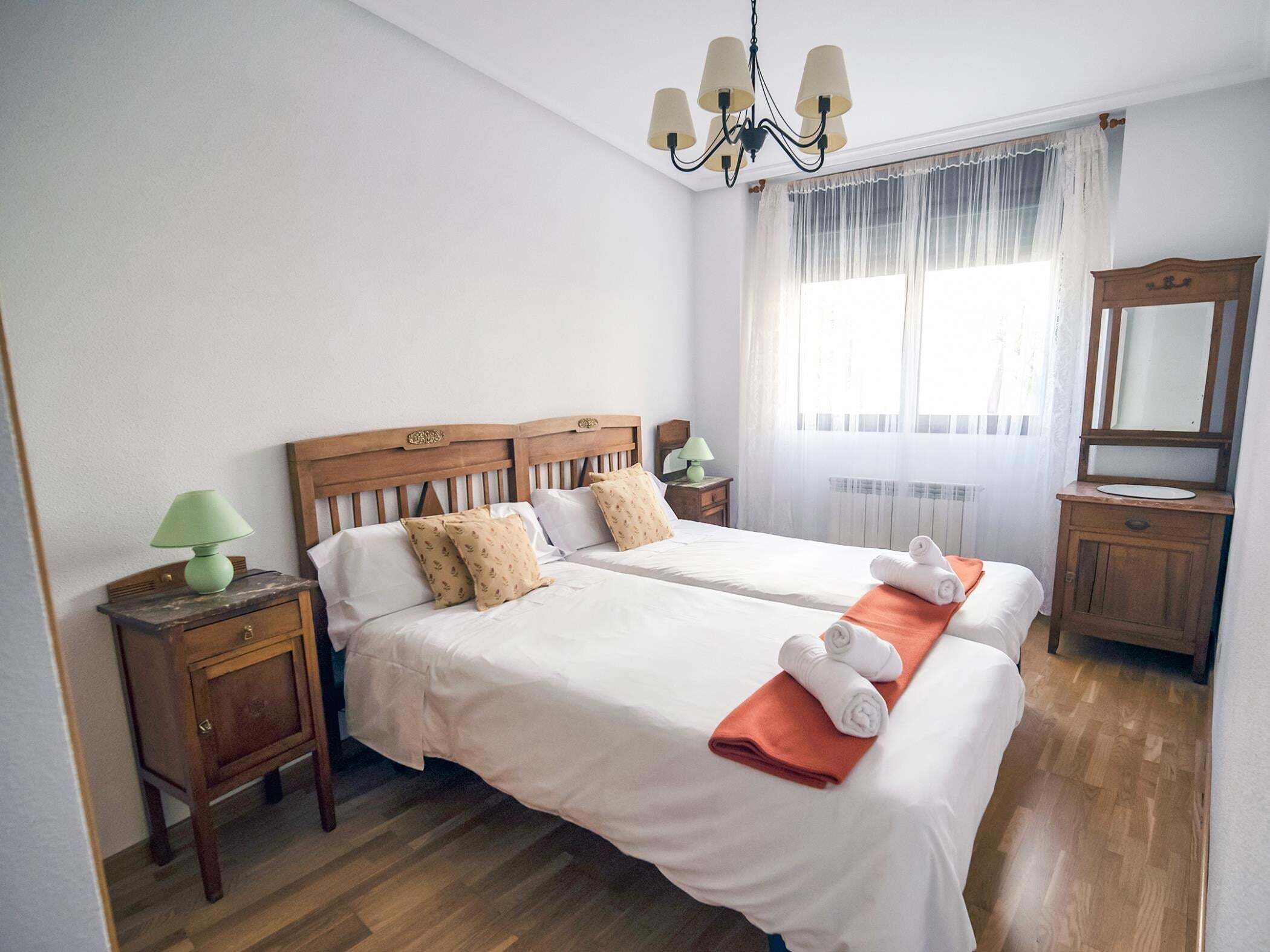 Una cama doble con sábanas blancas, cabecera de madera, mesita de noche con lámpara, espejo y ventana con cortinas traslúcidas.