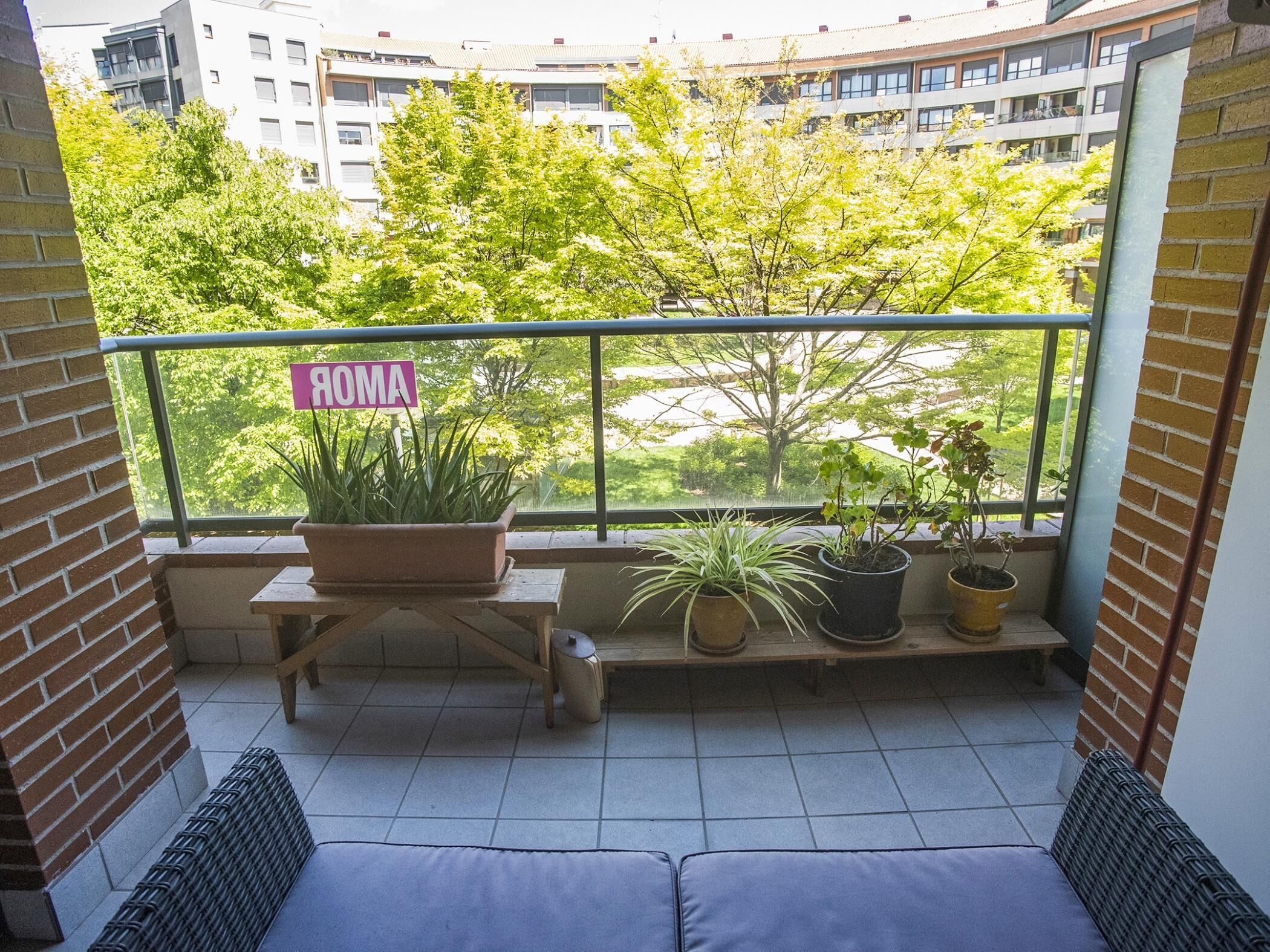 A balcony with a sign reading 'ROMA,' potted plants, a wooden bench, and a view of buildings and trees.