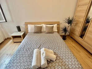Ein ordentlich bezogenes Bett mit einem geometrisch gemusterten Überwurf, zwei Kissen und zwei zusammengerollten Handtüchern.