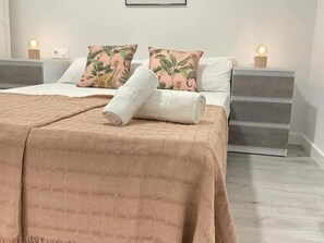 Una cama bien hecha con una manta rosa y dos almohadas, una mesita de noche con una lámpara y un cuadro colgado en la pared.