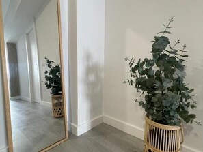 Un pasillo con un espejo que refleja una planta en una cesta de mimbre, una puerta y un suelo de color claro.