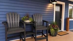 Dos sillas Adirondack en una terraza de madera, una planta en maceta con flores y una puerta con ventana.