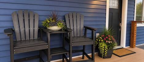 Dos sillas Adirondack en una terraza de madera, una planta en maceta con flores y una puerta con ventana.
