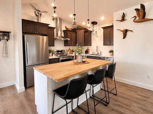 Une cuisine moderne dotée d’un îlot central en bois, d’appareils électroménagers en acier inoxydable, de luminaires suspendus et de sculptures d’oiseaux en bois fixées au mur.