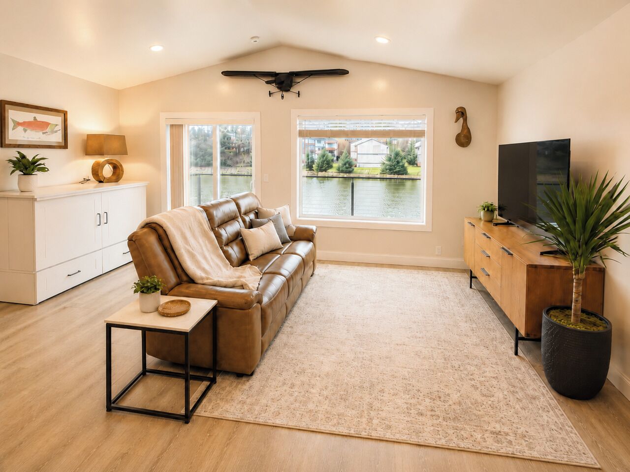 A living room with a leather sofa, a coffee table, a TV, a cabinet, and a large window with a view of a body of water.