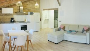 Una cucina moderna con elettrodomestici bianchi, una zona pranzo con tavolo e sedie bianche, e un soggiorno con divano e poggiapiedi bianchi.