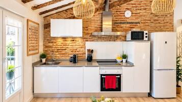 A modern kitchen with exposed brick walls, wooden beams, a stainless steel range hood, a microwave, a refrigerator, and a dining area with a table and chairs.