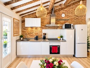 A modern kitchen with exposed brick walls, wooden beams, a stainless steel range hood, a microwave, a refrigerator, and a dining area with a table and chairs. - Loft Laurel acogedor (Logroño)