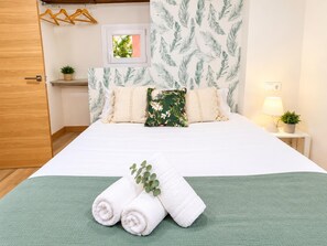 A neatly made bed with white linens, a green throw blanket, and rolled white towels. There is a small bedside table with a lamp and a potted plant. - El Rincón del Marqués by El Rincon (Logroño)