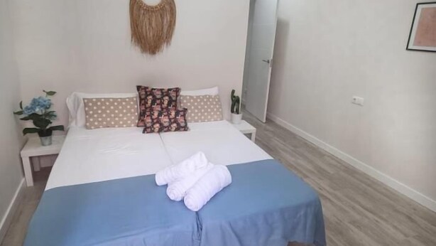 Een slaapkamer met een bed, kussens, een blauwe spreidek, wanddecoratie van macramé, een nachtkastje met een plant en een deur.