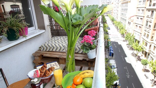 En balkon med spiseplads, komplet med ristet brød, en kop, en skål med frugt og en juiceflaske. Der er krukker med planter og et vindue med udsigt til en gade og bygninger.