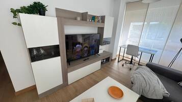 Un salon moderne doté d’un téléviseur à écran plat, d’une table basse blanche, d’un canapé gris et d’une grande fenêtre avec des stores.