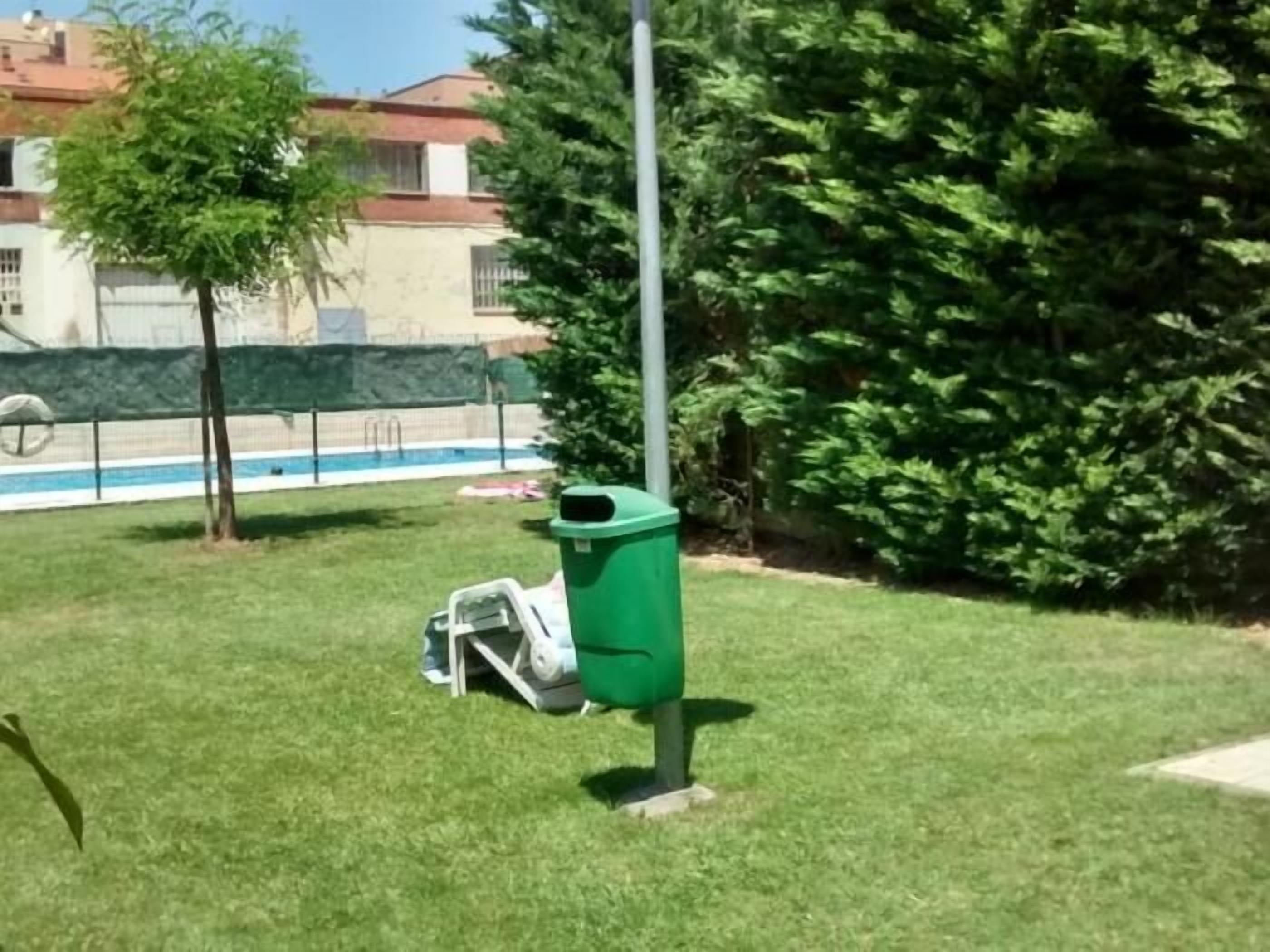 Uma lixeira verde em área gramada, com piscina e árvores ao fundo.