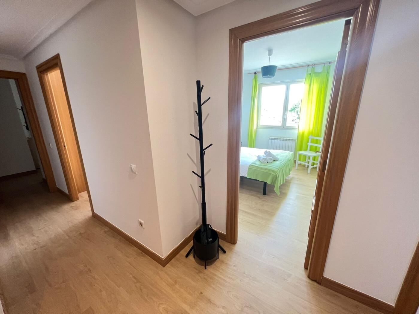 A hallway with a coat rack, a bed with a green bedspread, and a window with curtains.