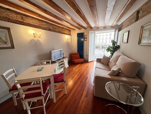 Een kamer met een houten plafond, een bank, een televisie, een eettafel met stoelen en een raam met uitzicht op gebouwen.
