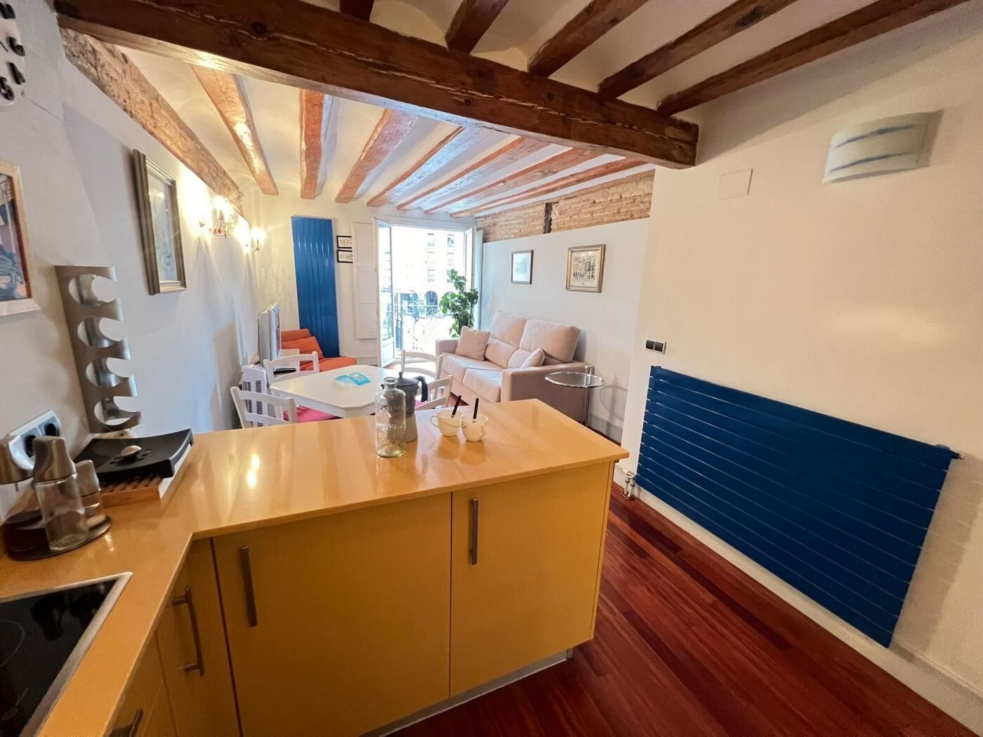 Una cocina compacta con una isla central, un comedor con mesa y sillas, y una sala de estar con sofá y radiador.