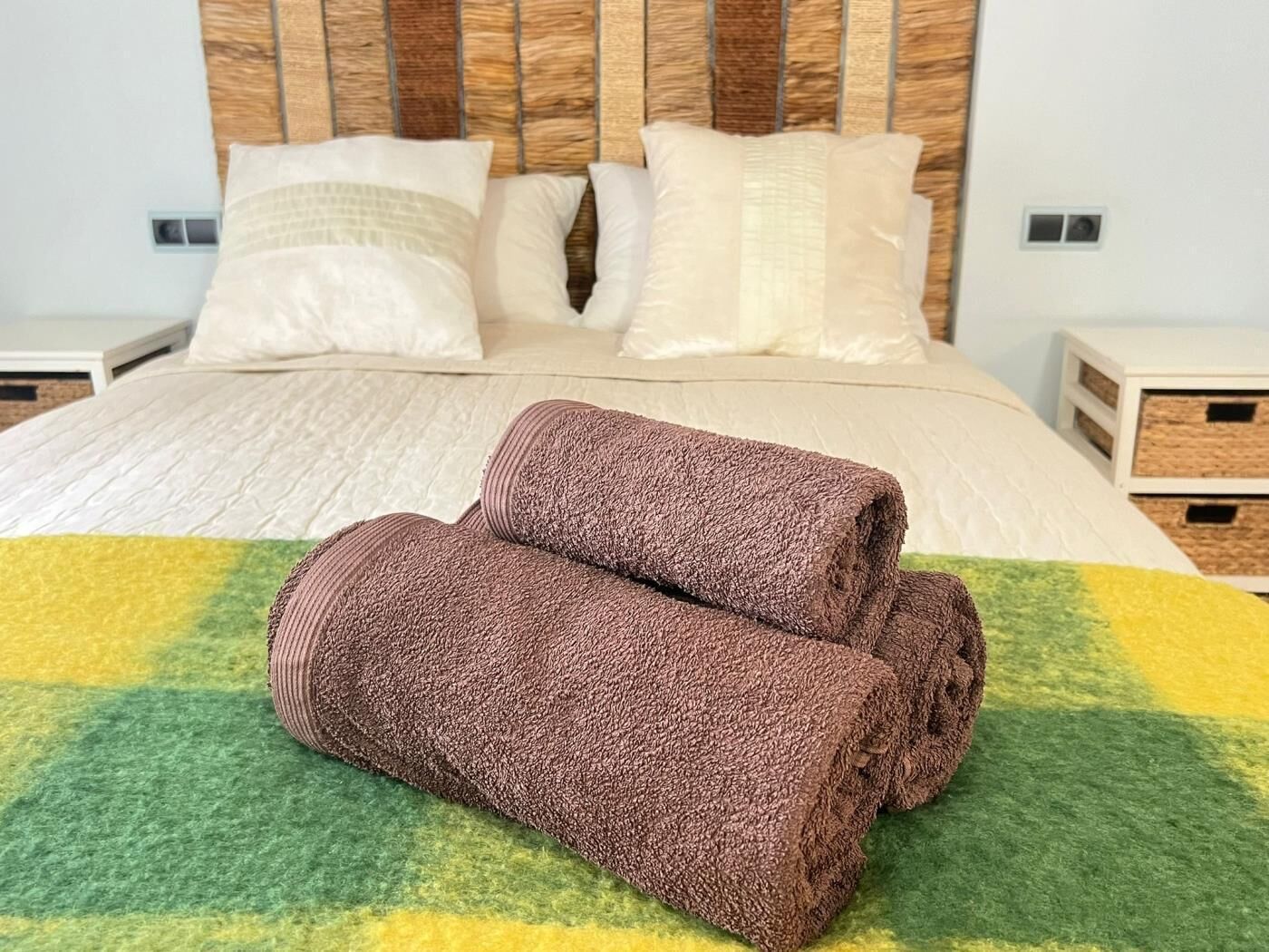 A neatly made bed with a green and yellow blanket, two rolled towels, and a wooden headboard.