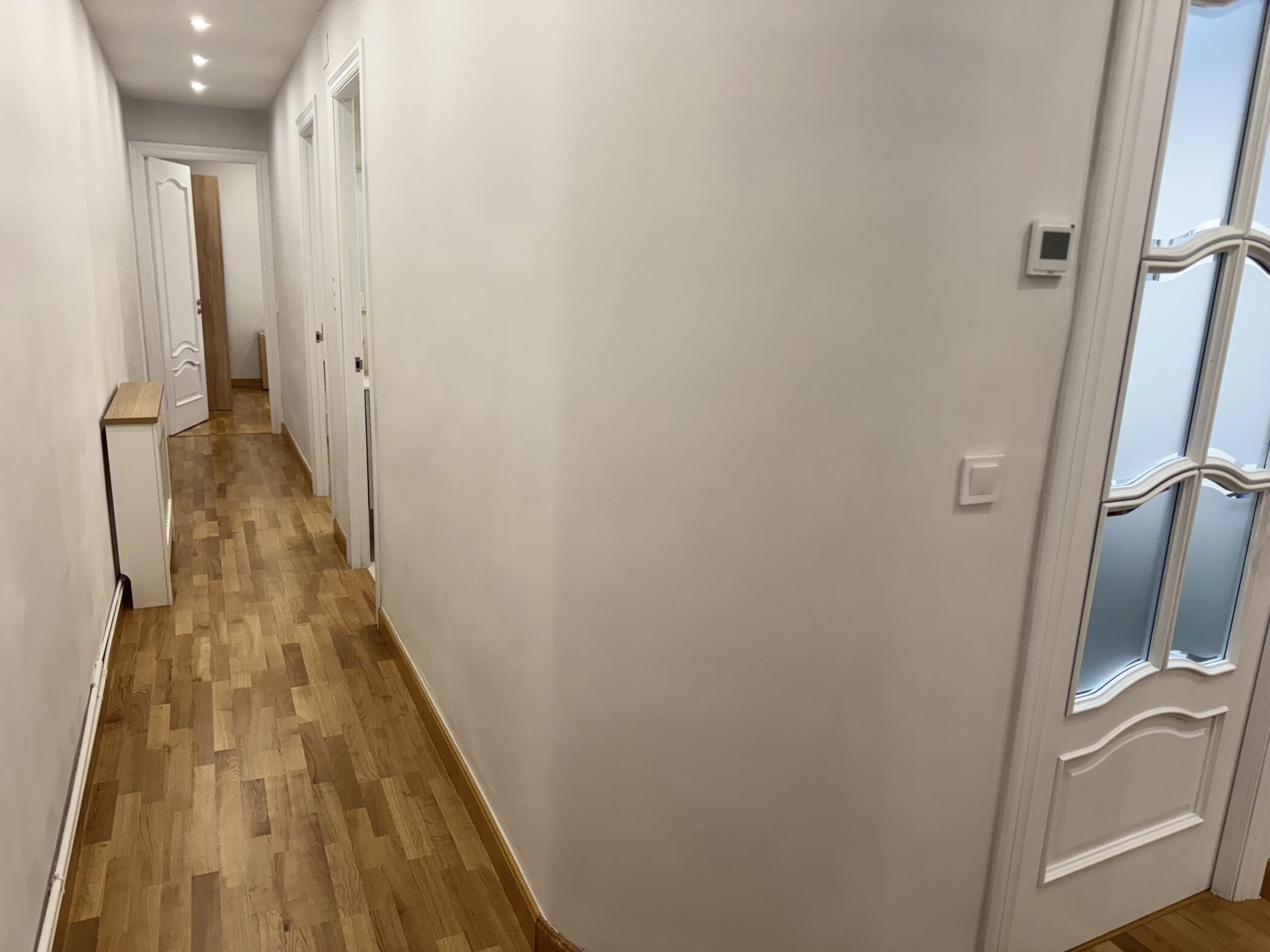 Een gang met houten vloer, witte muren en een deur met een decoratief glasraam.
