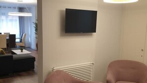 En moderne stue med et fladskærms-tv, to pink lænestole, en hvid radiator samt et spiseområde med bord og stole.