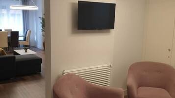 A modern living room with a flat-screen TV, two pink armchairs, a white radiator, and a dining area with a table and chairs.