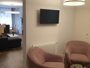 Ein modernes Wohnzimmer mit einem Flachbildfernseher, zwei rosafarbenen Sesseln, einem weißen Heizkörper und einem Essbereich mit Tisch und Stühlen.