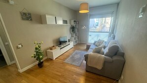 A living room with a grey sofa, a TV on a white stand, a potted plant, and a window with a view of buildings. - Santiago Tower Views (Logroño)