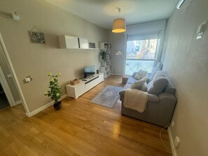 Una sala de estar con un sofá gris, un televisor sobre un mueble blanco, una planta en maceta y una ventana con vistas a edificios.