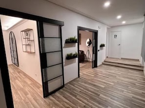 Un pasillo con suelo de madera, una puerta corredera, un estante empotrado con plantas y una puerta con un cartel.