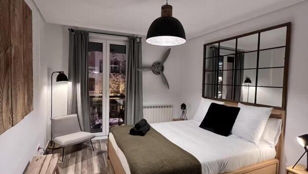 Un dormitorio con cama, mesita de noche, lámpara y vistas al exterior.