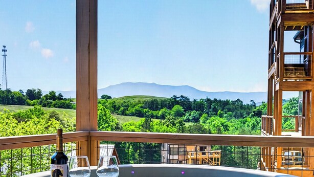Ein Whirlpool mit einer Flasche und Gläsern, eine Holzterrasse und Blick auf Grünflächen und Berge.