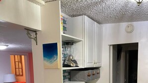 Eine kleine Küche mit weißen Schränken, einer Arbeitsplatte und einer Spritzschutzwand. Ein Bild hängt an der Wand und eine Deckenleuchte ist angebracht.