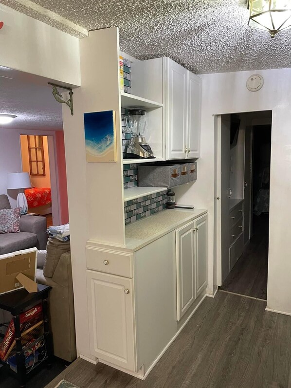 A compact kitchen with white cabinets, a countertop, and a backsplash. There is a painting on the wall and a ceiling light fixture.