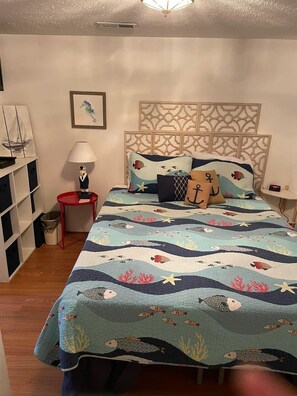 Un dormitorio con cama, mesita de noche, lámpara, estante con libros y un reloj de pared.