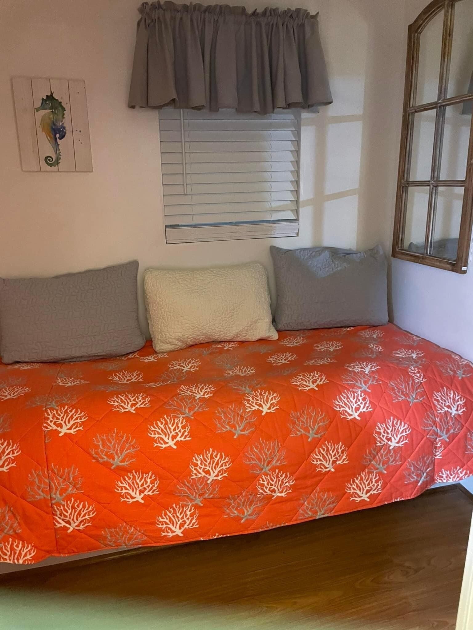 Ein Bett mit roter Decke, zwei Kissen, ein Fenster mit Jalousien und einer Wanddekoration.
