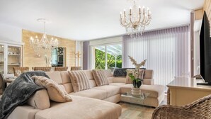 A modern living room with a sectional sofa, a glass coffee table, a chandelier, and a television. - Gîtes de France® - La Margautine (VEULETTES SUR MER)