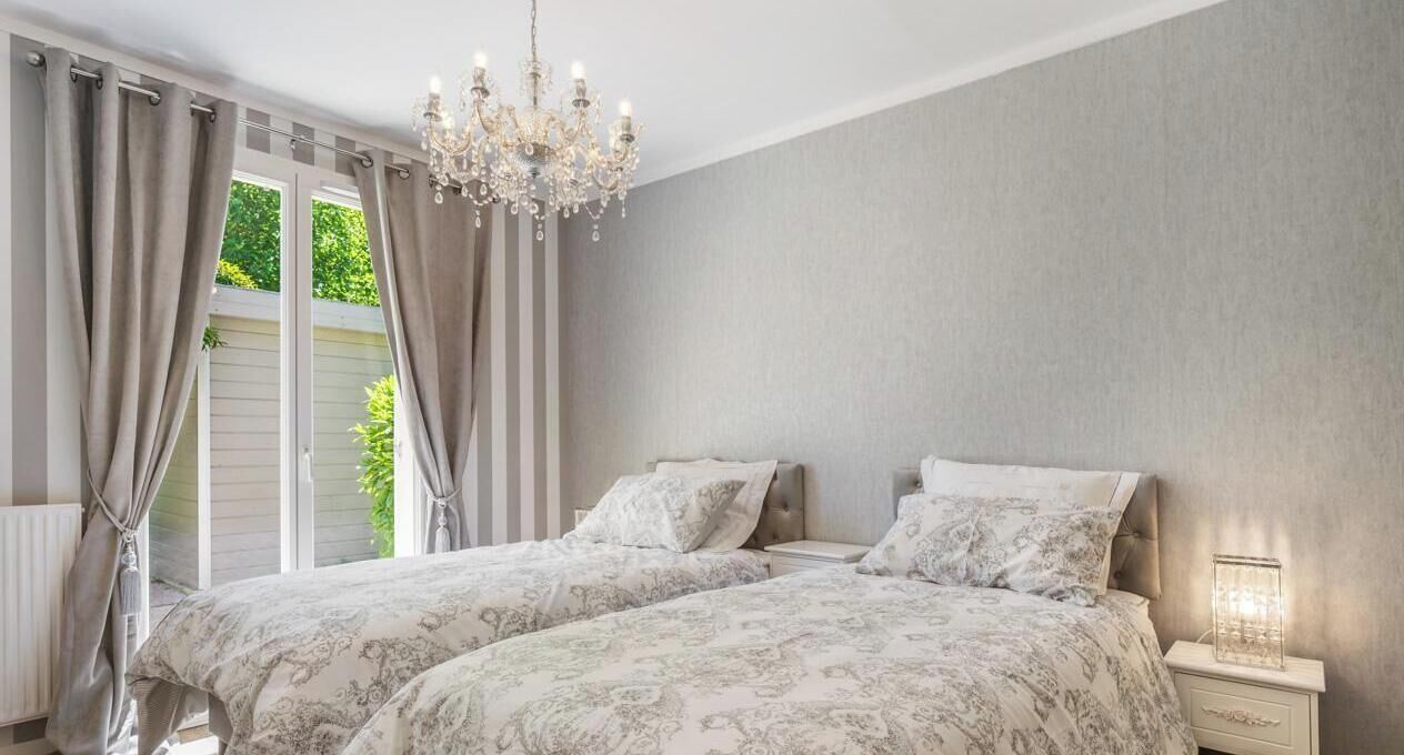 Een slaapkamer met twee bedden, een kroonluchter, een nachtkastje met een lamp en uitzicht op groen buiten.