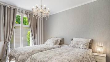 Een slaapkamer met twee bedden, een kroonluchter, een nachtkastje met een lamp en uitzicht op groen buiten.
