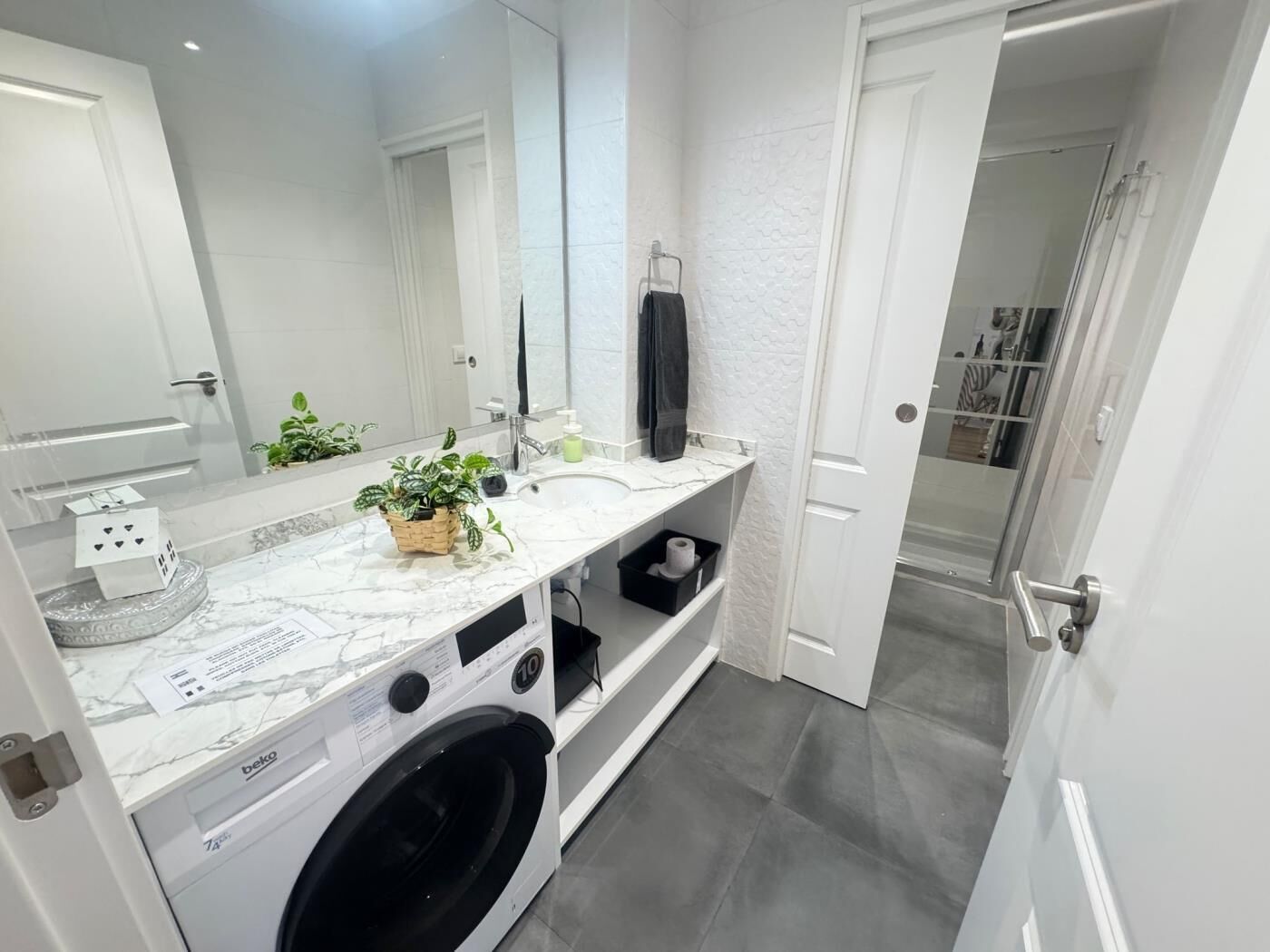A modern bathroom with a white marble sink, a large mirror, a washing machine, and a shower area.